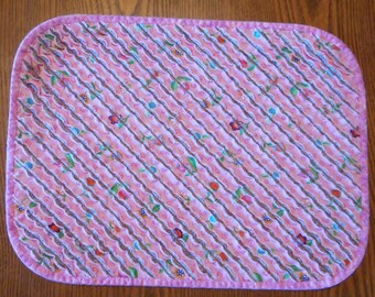 May include: A pink and white quilted table runner with a scalloped edge and a floral pattern. The runner has a pink border.