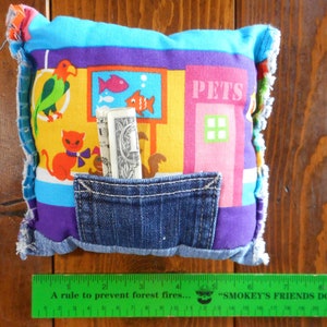Op de afbeelding: Een kleurrijk, handgemaakt kussen met een spijkerbroekzak. Het kussen heeft een patchwork-ontwerp met cartoon dieren, een aquarium en het woord "PETS". Het kussen heeft onderaan een groene liniaal met de tekst "A rule to prevent forest fires..."