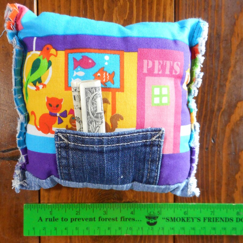 Op de afbeelding: Een kleurrijk, handgemaakt kussen met een spijkerbroekzak. Het kussen heeft een patchwork-ontwerp met cartoon dieren, een aquarium en het woord "PETS". Het kussen heeft onderaan een groene liniaal met de tekst "A rule to prevent forest fires..."