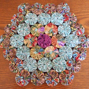May include: A circular fabric mat constructed from numerous gathered fabric yo-yos in a variety of patterns and colors, including shades of blue, green, red, yellow, and a single solid purple yo-yo positioned near the center of the arrangement.