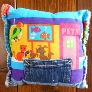Op de afbeelding: Een kleurrijk, handgemaakt kussen met een dierenwinkelontwerp. Het kussen heeft een spijkerbroekzak en is versierd met cartoon dieren, waaronder een papegaai, kat, hond en vissen. Het woord "PETS" staat op het kussen.