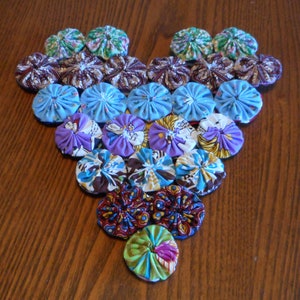 Puede incluir: Un arreglo en forma de corazón de flores de tela en varios colores, incluyendo azul, morado, verde y marrón. Las flores están dispuestas en filas, con las flores más grandes en la parte inferior y las flores más pequeñas en la parte superior.