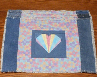 May include: A blue and pink patchwork table runner with a heart-shaped design in the center. The heart is made of fabric with a rainbow pattern. The runner has a frayed edge.