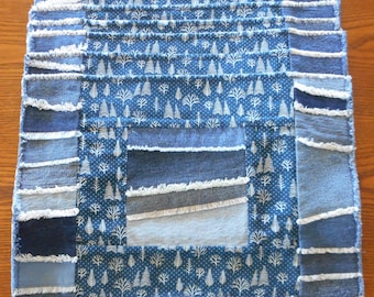 May include: A blue and white denim placemat with a patchwork design. The placemat is made of different pieces of denim fabric, some of which have been frayed. The placemat has a white pattern of small trees on a blue background.