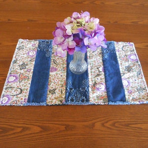May include: A blue and white patchwork table runner with a frayed edge. A bouquet of purple and white flowers in a clear glass vase sits on top of the runner.