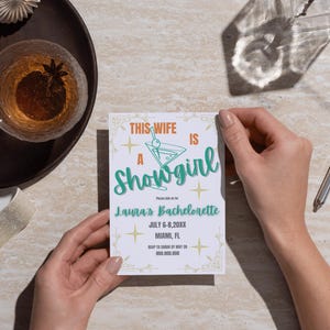 Puede incluir: Una tarjeta de invitación blanca con el texto "This Wife Is A Showgirl" en naranja y verde azulado. La tarjeta también incluye detalles para la despedida de soltera de Laura en Miami, FL. La tarjeta es sostenida por una persona.