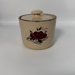 以下が含まれることがあります： ベージュ色の蓋付き陶器の瓶。瓶には、緑の葉と黒のアクセントが付いた2つの赤いリンゴの手描きのデザインが施されています。瓶の縁には濃い青色のトリムが付いています。