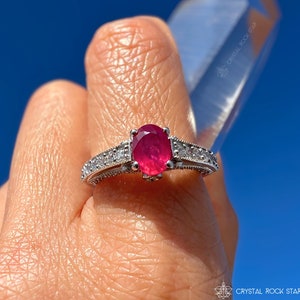 May include: A silver ring with a large oval ruby gemstone and small accent stones. The ring is held up against a bright blue sky. The ring's band has a detailed design.