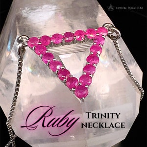 May include: A silver Trinity necklace with a triangular pendant set with vibrant pink rubies. The necklace has a delicate chain and is displayed on a clear, crystalline surface. The word "Ruby" is written in a cursive font.