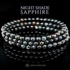 May include: Three stacked bracelets made of dark gray, spherical beads. The beads have a metallic sheen and reflect light, with some appearing blue or green. The text "NIGHT SHADE SAPPHIRE" is at the top, and "CRYSTAL ROCK STAR" is at the bottom.