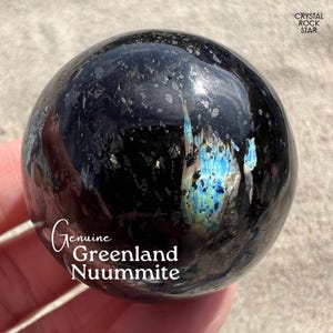 May include: A polished spherical Nuummite stone featuring a deep black base color with scattered metallic flecks and a prominent iridescent blue and gold flash. The text "Genuine Greenland Nuummite" and "CRYSTAL ROCK STAR" is printed on the image.