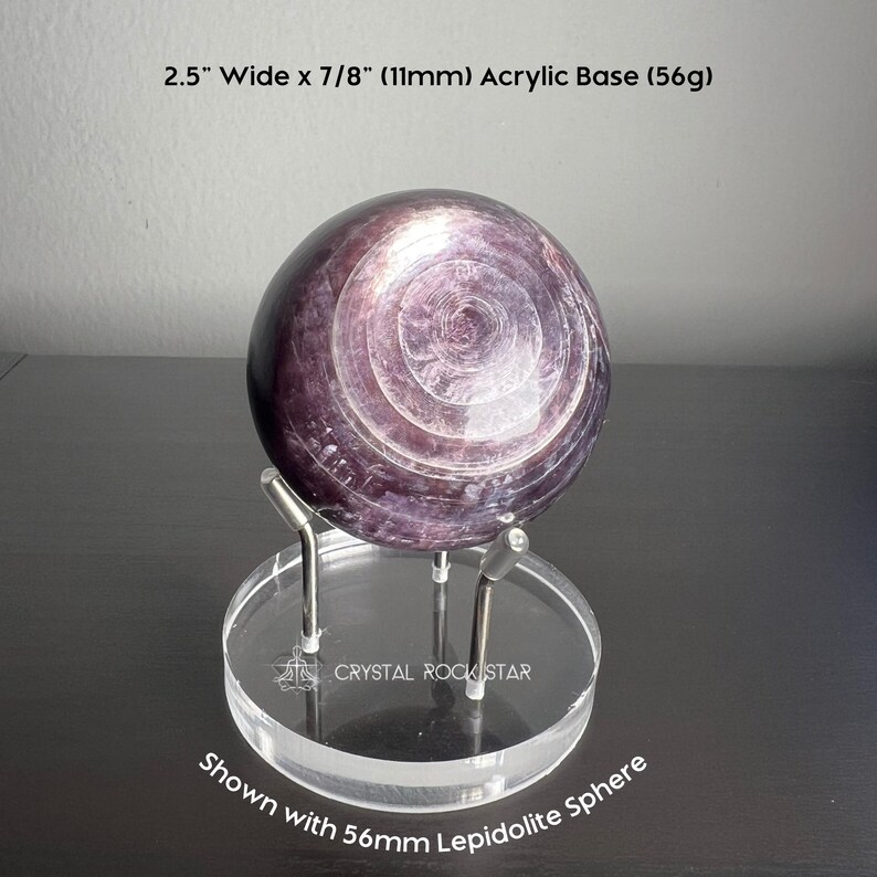 Puede incluir: Una esfera de lepidolita de color p&uacute;rpura oscuro con patrones circulares conc&eacute;ntricos sobre una base de acr&iacute;lico transparente con tres puntas de metal. La base mide 6,35 cm de ancho por 11 mm de alto y pesa 56g. El texto CRYSTAL ROCKSTAR aparece en la base.