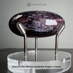 Puede incluir: Un cristal lingam de lepidolita de 68 mm con tonos p&uacute;rpuras profundos y oscuros, que presenta una forma ovalada lisa y pulida. El cristal se exhibe sobre un soporte circular de acr&iacute;lico transparente sostenido por tres puntas de metal plateado.