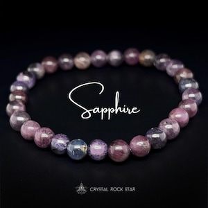 May include: A bracelet made of round, polished sapphire beads in shades of purple, pink, and blue. The word "Sapphire" is written in cursive script. The bracelet is displayed against a black background.