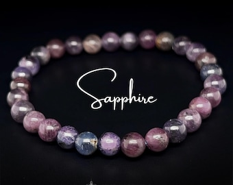 Puede incluir: Un brazalete de cuentas redondas y pulidas de zafiro en tonos morados, rosas y azules. La palabra "Sapphire" está escrita en cursiva. El brazalete se muestra sobre un fondo negro.