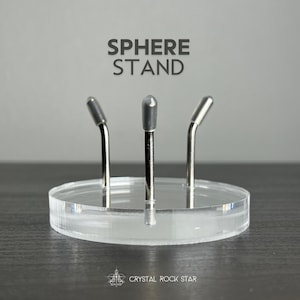 Puede incluir: Una base circular de acr&iacute;lico transparente con tres puntas met&aacute;licas verticales que tienen puntas protectoras grises. El texto SPHERE STAND est&aacute; impreso arriba y el texto CRYSTAL ROCK STAR est&aacute; impreso debajo del objeto en la base.