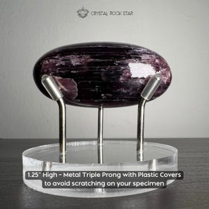 Puede incluir: Un esp&eacute;cimen de cristal p&uacute;rpura oscuro pulido de forma ovalada sobre una base circular de acr&iacute;lico transparente. El soporte tiene tres puntas met&aacute;licas con cubiertas de pl&aacute;stico y mide 3,175 cm de alto. El texto dice CRYSTAL ROCK STAR.