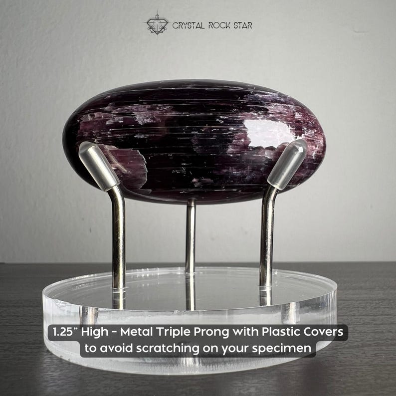 Puede incluir: Un esp&eacute;cimen de cristal p&uacute;rpura oscuro pulido de forma ovalada sobre una base circular de acr&iacute;lico transparente. El soporte tiene tres puntas met&aacute;licas con cubiertas de pl&aacute;stico y mide 3,175 cm de alto. El texto dice CRYSTAL ROCK STAR.
