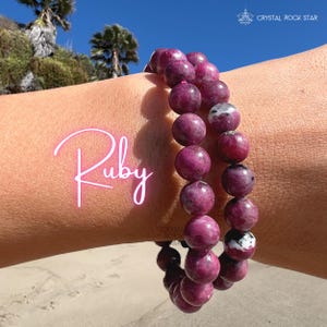 May include: A double-strand bracelet composed of round, polished ruby beads in varying shades of deep magenta and dark pink, featuring occasional white and black mineral inclusions. The word Ruby is written in pink script on the left side.