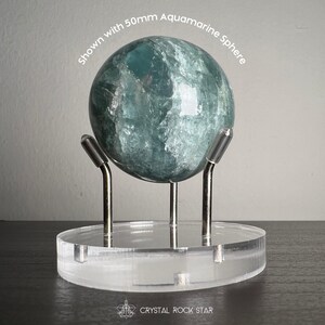 Puede incluir: Una esfera de aguamarina de 50 mm con textura cristalina azul verdosa transl&uacute;cida, sobre una base circular de acr&iacute;lico transparente con tres soportes met&aacute;licos plateados. Se lee el texto "Shown with 50mm Aquamarine Sphere" y "CRYSTAL ROCK STAR".