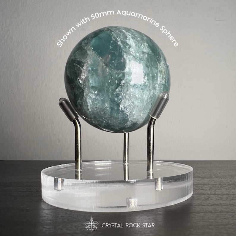 Puede incluir: Una esfera de aguamarina de 50 mm con textura cristalina azul verdosa transl&uacute;cida, sobre una base circular de acr&iacute;lico transparente con tres soportes met&aacute;licos plateados. Se lee el texto "Shown with 50mm Aquamarine Sphere" y "CRYSTAL ROCK STAR".