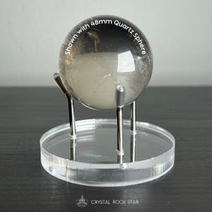 Puede incluir: Una esfera de cuarzo transparente de 48 mm con inclusiones oscuras sobre un soporte de tr&iacute;pode de metal plateado. El soporte est&aacute; montado sobre una base circular de acr&iacute;lico transparente. El texto "Shown with 48mm Quartz Sphere" aparece impreso.