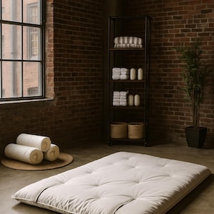 May include: A white, tufted futon mattress with a neutral-toned cover, placed on a concrete floor. Rolled white pillows rest on a round woven mat. A black metal shelf holds towels and baskets, and a potted plant is in the background.