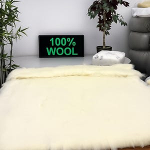 May include: A cream-colored wool rug or blanket is displayed on a wooden surface. The text "100% WOOL" is visible on a black sign. The background includes plants and a stack of pillows.