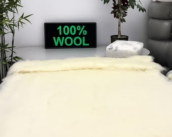 May include: A cream-colored wool rug or blanket is displayed on a wooden surface. The text "100% WOOL" is visible on a black sign. The background includes plants and a stack of pillows.