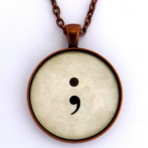 Puede incluir: Un collar de cadena de cobre con un colgante redondo que presenta un punto y coma negro sobre un fondo blanco.