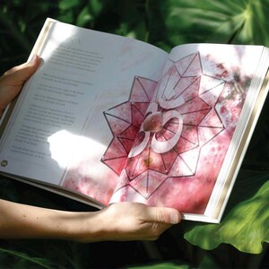 May include: An open book with a pink and white geometric design and the symbol "Om" in the center. The book is held open by a person's hands, with text visible on the left page. The background is blurred green foliage.