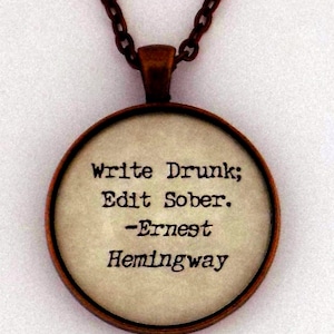 Puede incluir: Un collar redondo de color cobre con un colgante de vidrio. El colgante tiene un fondo blanco con texto negro que dice "Write Drunk; Edit Sober. -Ernest Hemingway"