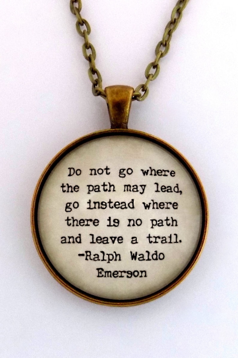 May include: A gold chain necklace with a round pendant featuring a quote by Ralph Waldo Emerson: "Do not go where the path may lead, go instead where there is no path and leave a trail."