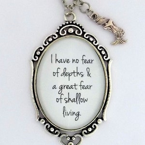May include: Silver necklace with a pendant featuring a quote: "I have no fear of depths & a great fear of shallow living." The pendant is in the shape of an oval frame with a heart at the bottom. A small mermaid charm hangs from the necklace.