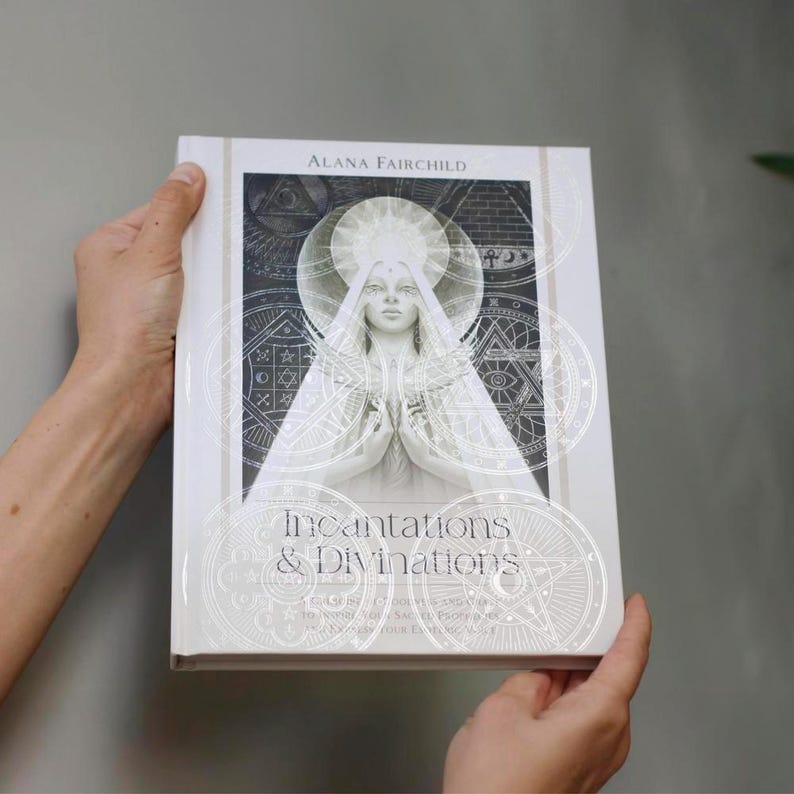 May include: A white book titled "Incantations & Divinations" by Alana Fairchild. The cover features a detailed illustration of a woman with a halo, surrounded by geometric and celestial designs in silver. The book is held by hands.