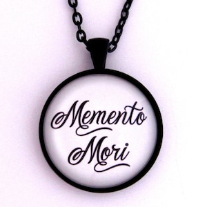 May include: A black chain necklace with a round pendant featuring the words "Memento Mori" in black script on a white background.
