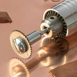 May include: A close-up of a silver rotary tool with a small, circular saw blade attached. The tool is resting on a copper surface.