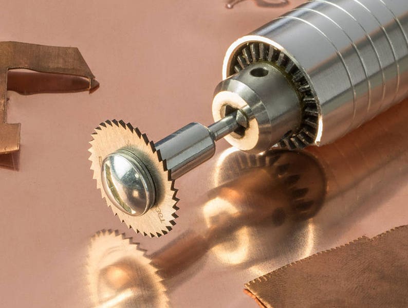 May include: A close-up of a silver rotary tool with a small, circular saw blade attached. The tool is resting on a copper surface.