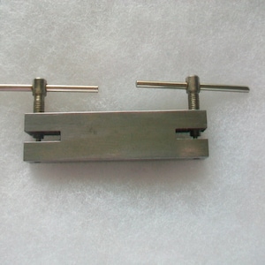 Peut inclure: Un outil de poinçonnage en métal argenté avec deux poignées réglables. L'outil a une base rectangulaire avec une forme découpée au centre.
