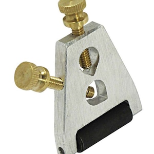 Puede incluir: Una herramienta de metal plateado con un rodillo de goma negro y dos pomos de latón. La herramienta se utiliza para afilar cuchillos.