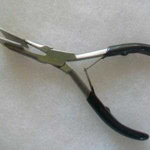 Puede incluir: Un par de alicates de metal plateado con mangos de plástico negro. Los alicates tienen un diseño único con una mandíbula plana y rectangular.