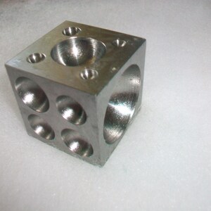 May include: A silver metal cube with multiple round depressions on each side. The cube has a smooth, polished finish.