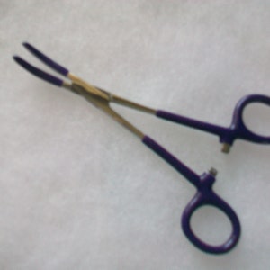 May include: Purple-handled surgical forceps with a silver metal body. The forceps have a locking mechanism and are used for grasping and holding small objects.