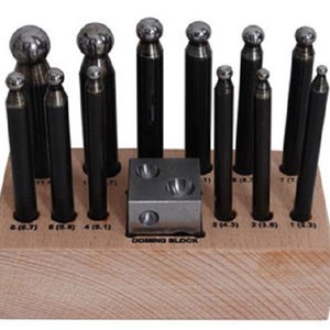 May include: A set of 10 metal dome punches in a wooden holder. The punches are arranged in order of size, with the smallest punch on the left and the largest punch on the right. The holder has a label that reads "DOMING BLOCK".