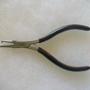 Op de afbeelding: Een tang van roestvrij staal met zwarte handvatten. De tang heeft een kleine, puntige punt en een grotere, platte kaak. De handvatten zijn gemaakt van een zacht, rubberachtig materiaal. De tang is gemarkeerd met "Stainless Steel Japan".