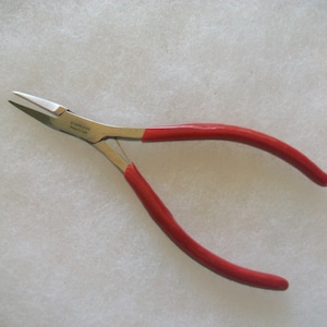 Puede incluir: Alicates de punta fina con mangos rojos y mordazas de acero inoxidable. Los alicates están hechos en Pakistán.