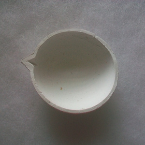 Puede incluir: Un crisol de cerámica blanca con un pico vertedor. El interior es liso y blanco, mientras que el borde exterior tiene un acento rojo. El crisol está diseñado para fundir sustancias a altas temperaturas, comúnmente utilizado en laboratorios o para la artesanía.