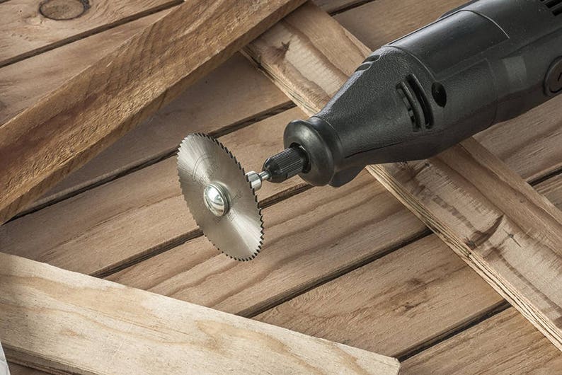May include: A black rotary tool with a small circular saw blade attached to the end. The tool is lying on a wooden surface.