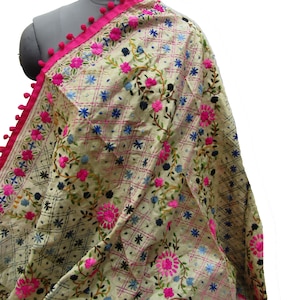 Könnte beinhalten: Cremefarbener bestickter Schal mit rosa Bordüre und Bommeln. Der Schal hat ein Blumenmuster in rosa, blau und schwarzem Faden. Das Design umfasst ein Gittermuster und florale Motive.