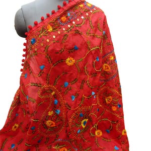 Op de afbeelding: Een felrode geborduurde sjaal met ingewikkelde bloemen- en paisley-patronen. De sjaal heeft gele, blauwe en oranje borduursels, samen met kleine pailletten. Rode pompons omlijsten de rand, wat een decoratief accent aan dit textiel toevoegt.
